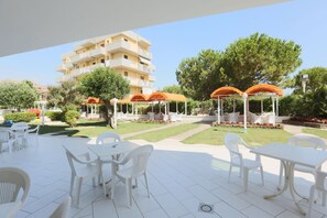 An outdoor patio area with white plastic tables and chairs, orange umbrellas, and a building in the background. - Hotel K2 (Ravenna)