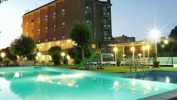 Una piscina con un tobogán verde, rodeada por un edificio de varios pisos y balcones, y un área cercada con mobiliario exterior.