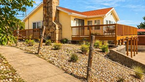 A two-story house with a stone chimney, a wooden deck, and a gravel driveway. - Buccaneer Bay #28 - Beachfront Bliss (Sunrise Beach)