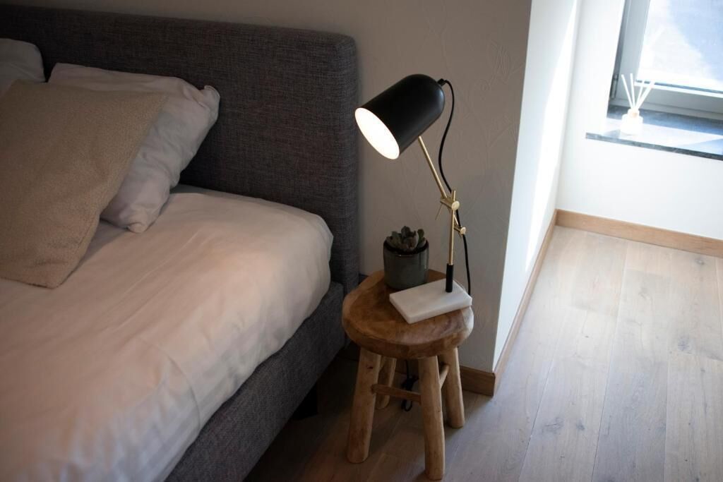 Ein Schlafzimmer mit einem Bett, einer Nachttischlampe, einem kleinen Hocker und einem Fenster.
