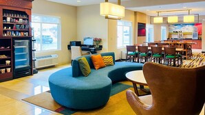 Un moderno hall de hotel con un sofá azul curvo, un sillón marrón, una zona de comedor con sillas verdes y una cocina al fondo.