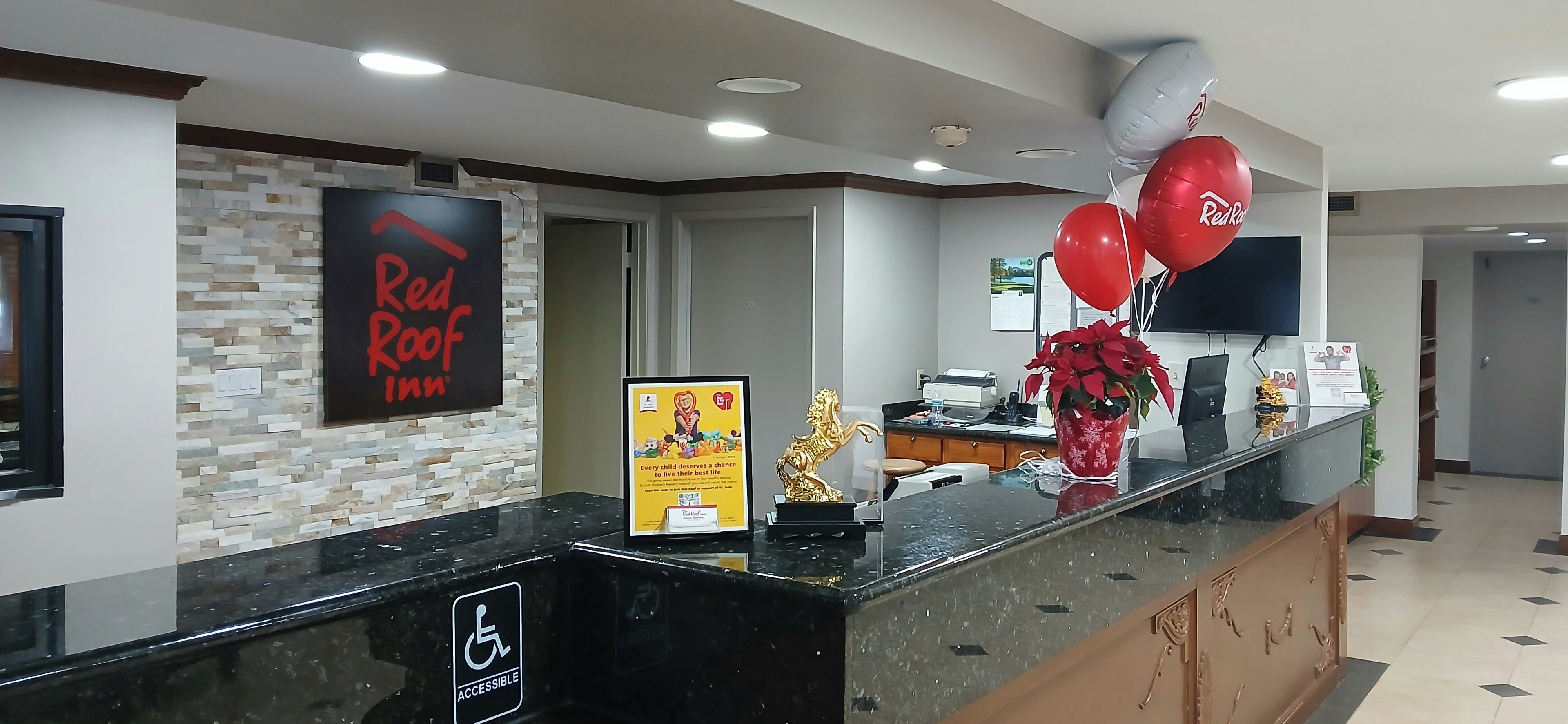 A hotel reception desk with a Red Roof Inn sign, a wheelchair accessibility sign, and a bulletin board with a children's poster.