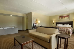 Habitación de hotel con sofá, una mesita, cama con colcha, mesita de noche, lámpara y un cuadro en la pared.