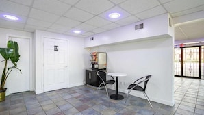Un pasillo con una puerta blanca, una máquina expendedora, una pequeña mesa con sillas y una planta en maceta.