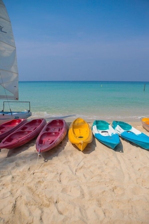 A row of colorful kayaks on a sandy beach with a sailboat in the background.