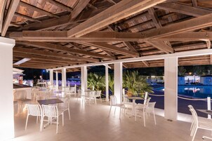 Un área de comedor al aire libre con sillas y mesas blancas, vigas de madera en el techo y vistas a una piscina y palmeras.