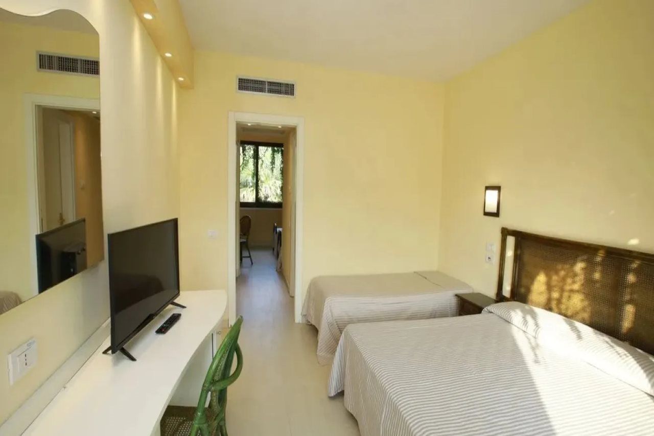 A hotel room with a television, two beds, a mirror, and a view of a corridor.