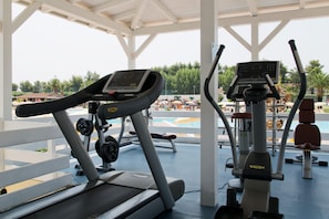 A covered outdoor gym area with treadmills and exercise equipment, overlooking a poolside with umbrellas and lounge chairs. - Futura Club Porto Kaleo (Cutro)