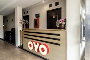 Un mostrador de recepción con detalles de madera y el logo 'OYO'. Hay dos arreglos florales sobre el mostrador, y una puerta con el número '005' en la parte superior.