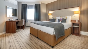 A hotel room with a large bed, a desk, and a chair. There is a window with curtains, a wall-mounted air conditioner, and a headboard with two lamps. - Garner Hotel Preston Samlesbury (Preston)