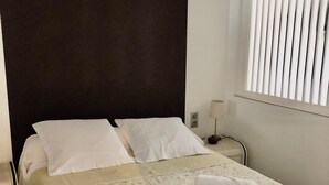 Et hotelværelse med seng, natbord, lampe og et vindue med persienner.