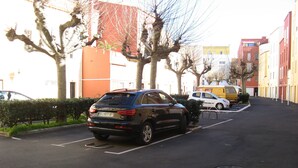 En parkeringsplads med en sort SUV, en gul varevogn og en hvid bil. Træer står langs vejen, og bygninger med altaner ses i baggrunden.