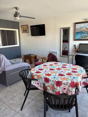 Een woonkamer met een bank, een tafel met bloemen tafelkleed, een televisie en een plafondventilator.
