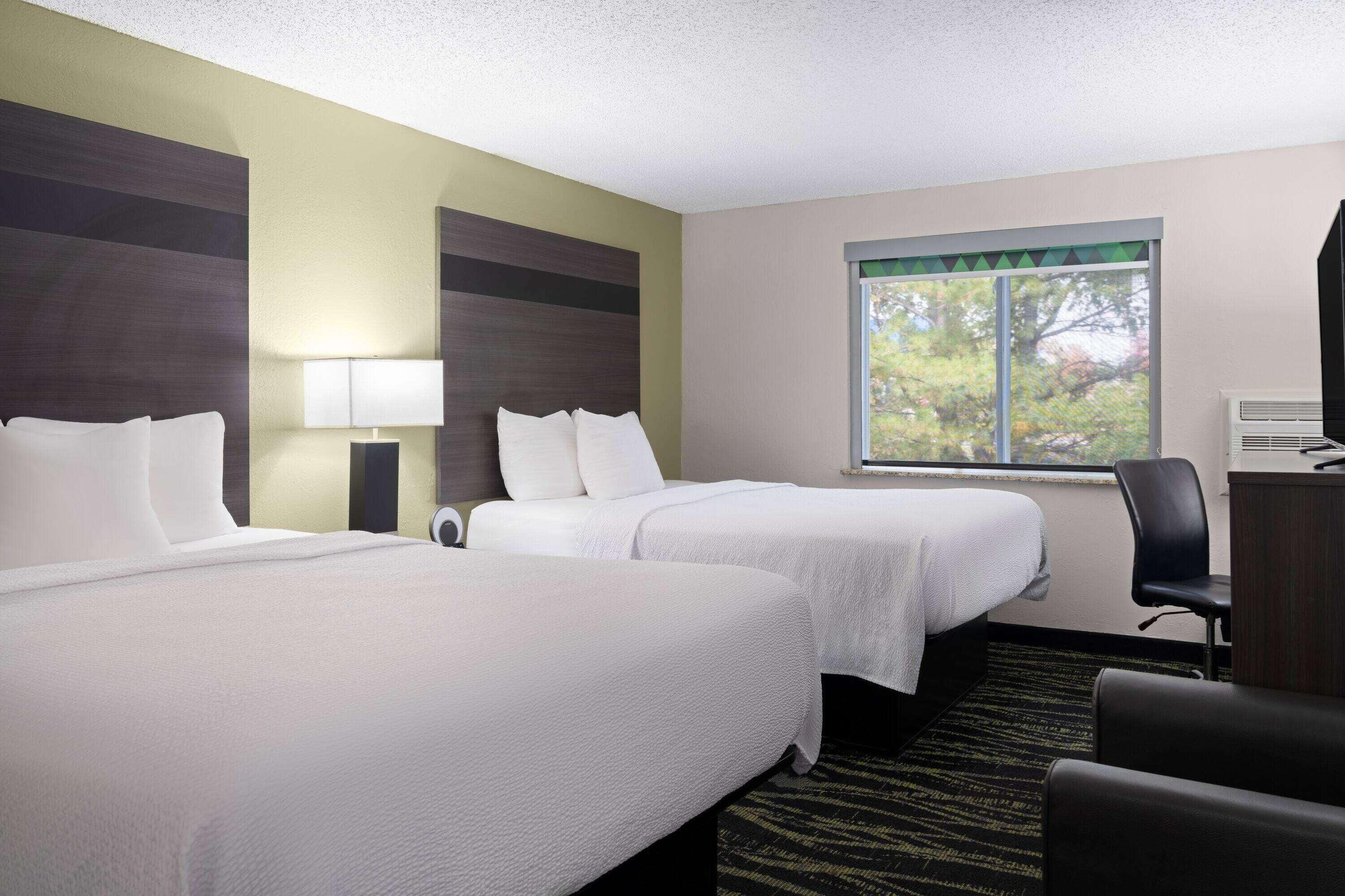 A hotel room with two beds, a desk, a chair, a lamp, and a window with a view of trees.
