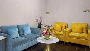 Ein Zimmer mit einem blauen Sofa, zwei gelben Sessel, einem runden Tisch mit Blumenvase und einem kleinen Regal mit Dekoration.
