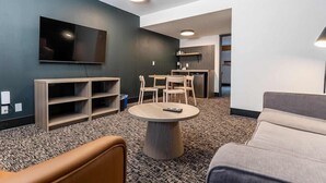 Un salon moderne avec un téléviseur à écran plat, une table basse ronde, un canapé et un coin repas comprenant des chaises et une petite cuisine en arrière-plan.