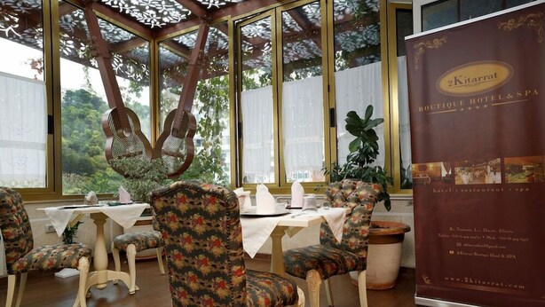 A dining area with floral upholstered chairs, a table with a white tablecloth, and a large window with a view of greenery. A promotional banner for '2Kitarat Boutique Hotel & Spa' is visible. - 2 Kitarrat Boutique Hotel and Spa (Durrës)