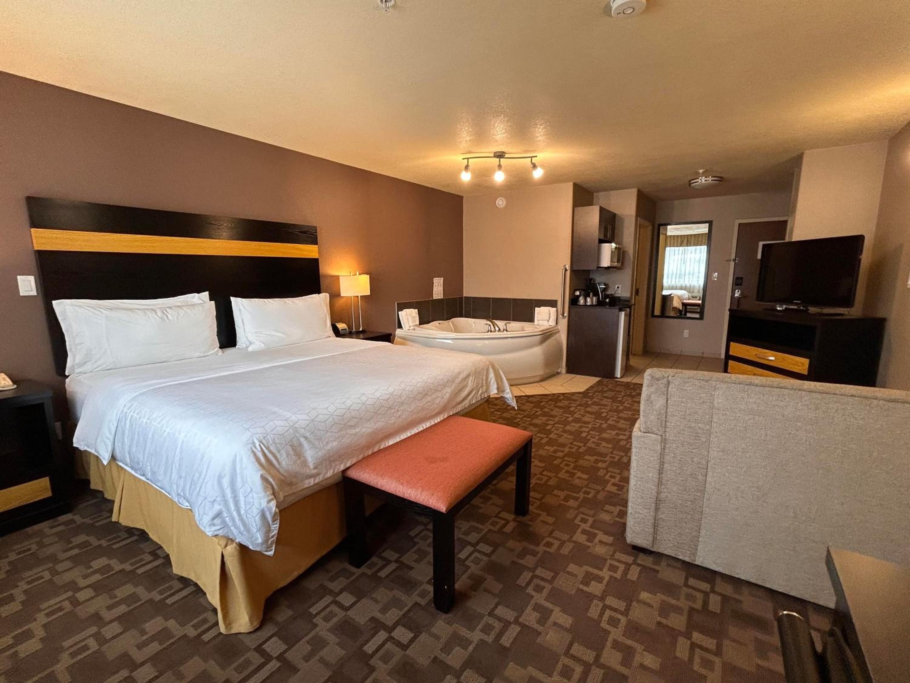 A hotel room with a large bed, a bench, a bathtub, a television, and a kitchenette.