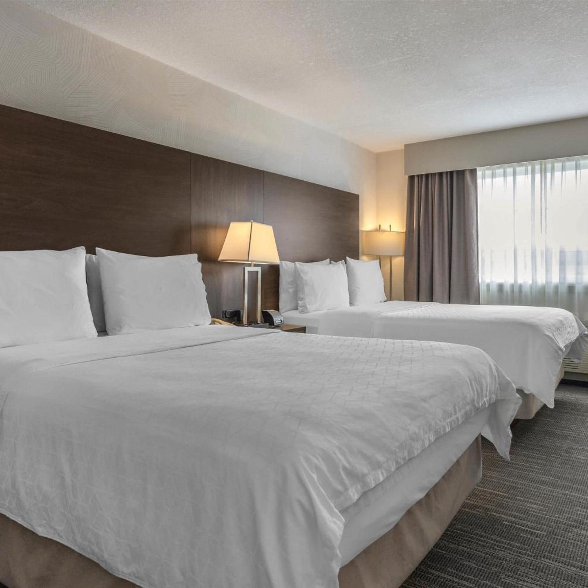 A hotel room with two beds, a nightstand, a lamp, and a window with curtains.