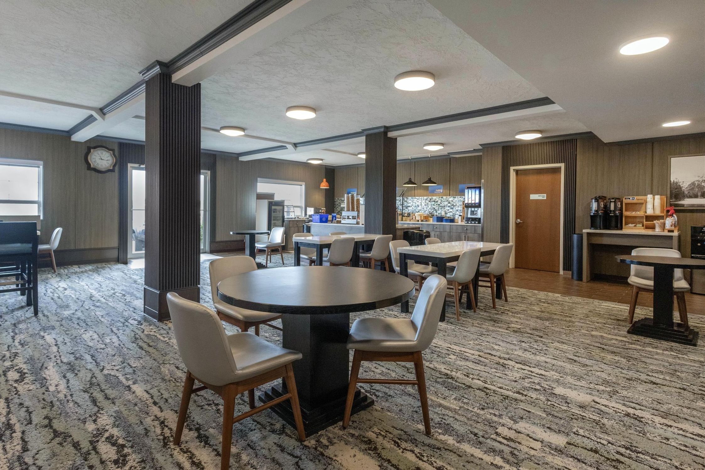 Un hall d’hôtel avec une table ronde, des chaises, un coin bar et une horloge au mur.