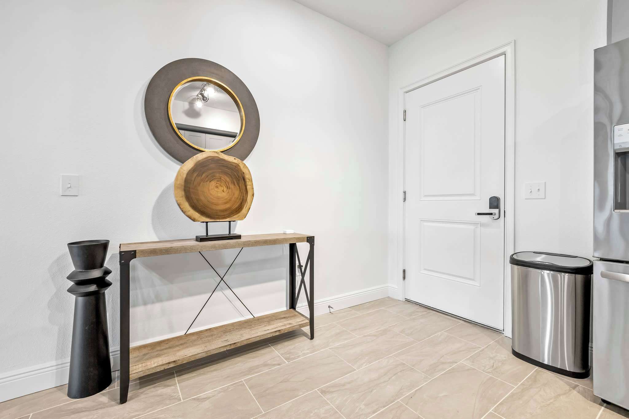 Une cuisine moderne avec une porte blanche, une poubelle en acier inoxydable, une console en bois avec un miroir et un vase noir.