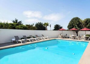 Ein Swimmingpool mit Liegestühlen und Sonnenschirmen, umgeben von einer weißen Mauer und viel Grün.