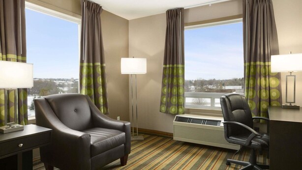 Una habitación de hotel con un sillón de cuero, un escritorio con lámpara, un pupitre con lámpara, un ventanal con cortinas y un radiador.