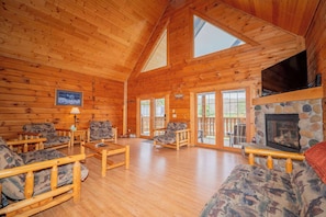 A log cabin interior with wooden walls, a stone fireplace, and a large window.