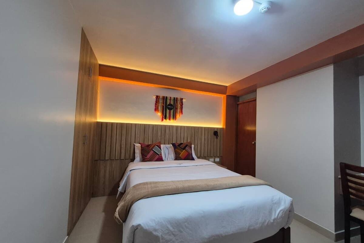 Quarto de hotel com cama, escrivaninha e luminária fixada na parede.