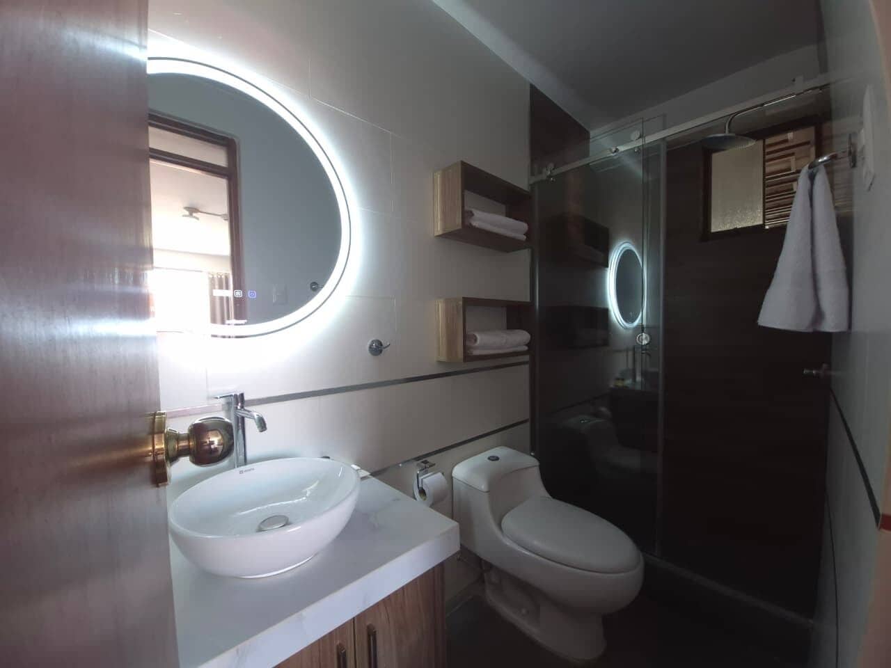 Un baño con un espejo redondo, dos lavamanos, un inodoro y una cabina de ducha de vidrio.