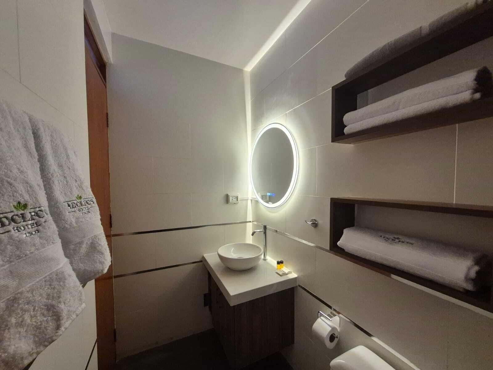 Un baño moderno con un espejo redondo, un lavamanos y un inodoro. Hay toallas en un estante y otra toalla colgada en la pared.