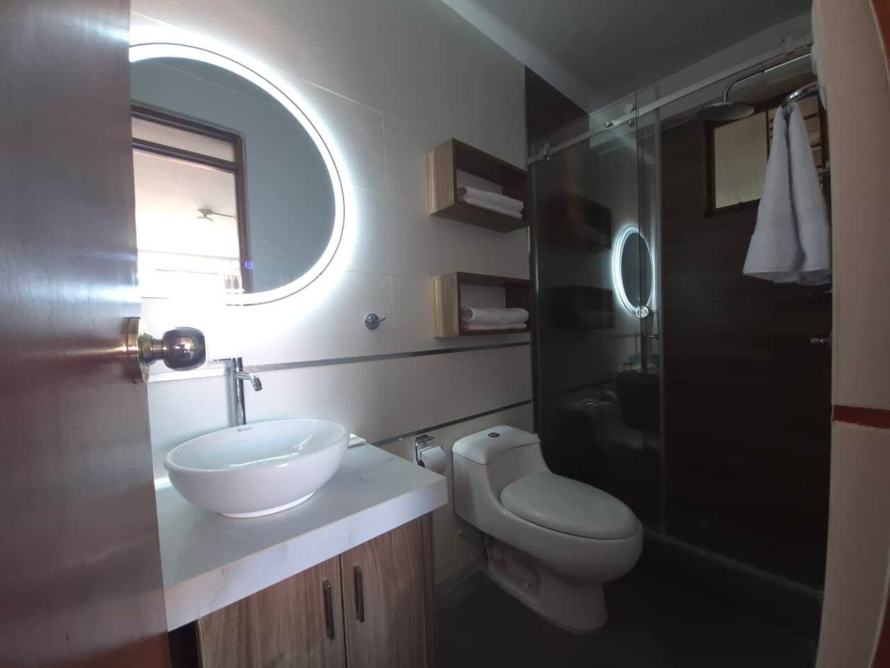 Un baño con un espejo redondo, un lavabo blanco, un inodoro y una cabina de ducha de vidrio.