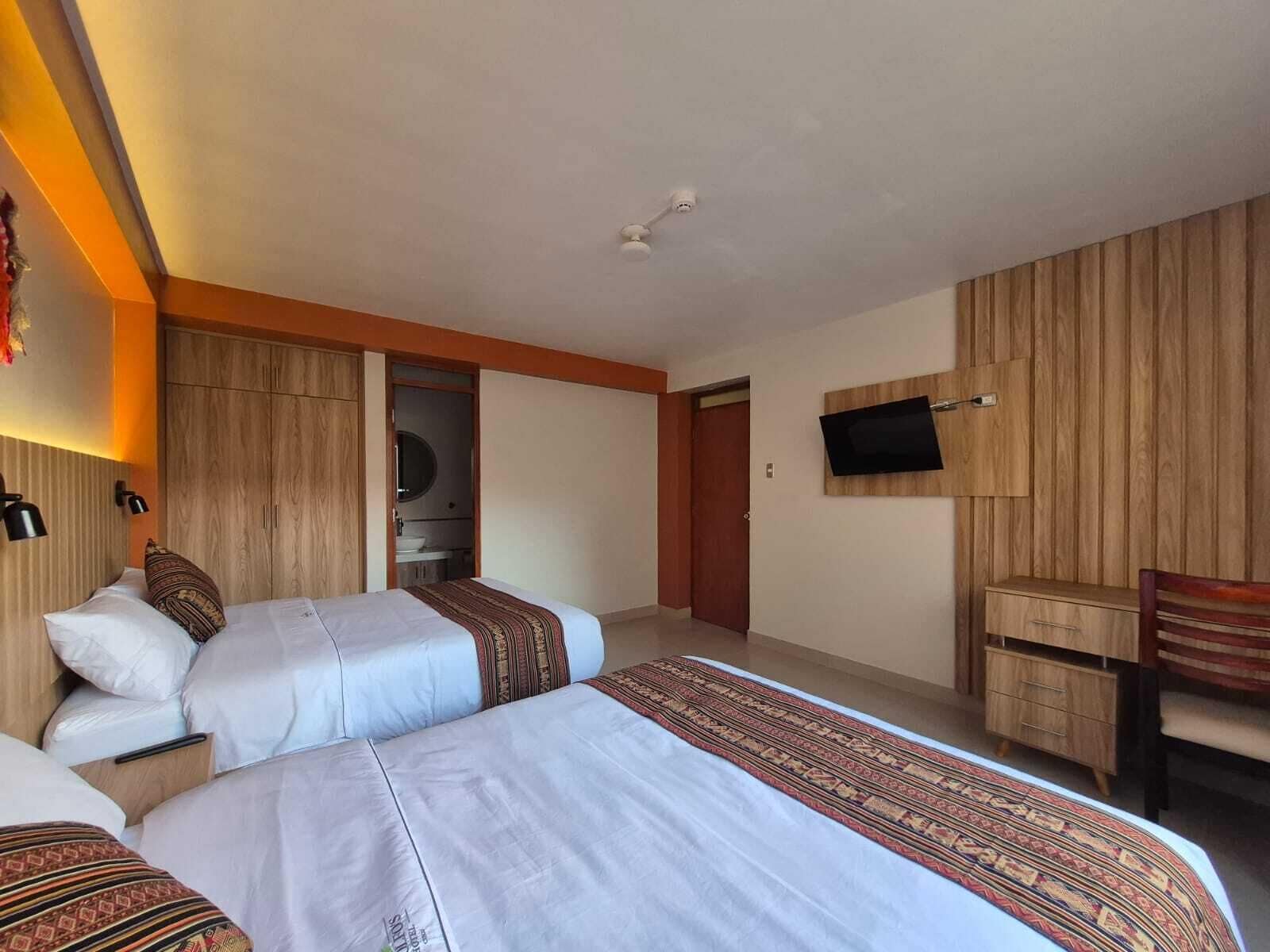 Een hotelkamer met twee bedden, een aan de muur bevestigde televisie, een houten garderobe en een klein bureau met lades.