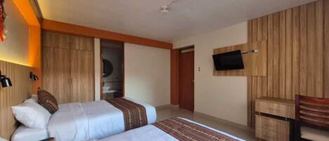 Een hotelkamer met twee bedden, een aan de muur bevestigde televisie, een houten garderobe en een klein bureau met lades.