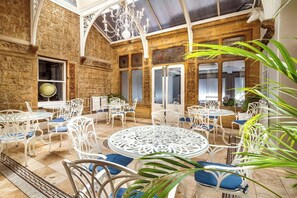 A spacious outdoor seating area with white metal chairs and tables, surrounded by stone walls and large windows. - The Shrubbery Hotel (Ilminster)