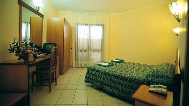 Una camera d'albergo con un letto, una scrivania, una sedia, uno specchio, un comodino e una finestra con tende.
