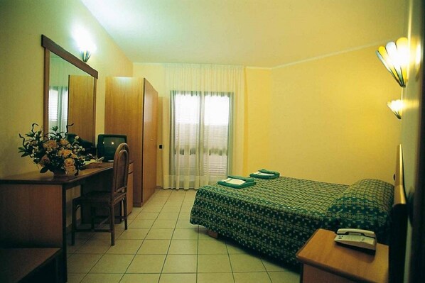 A hotel room with a bed, a desk, a chair, a mirror, a bedside table, and a window with curtains. - Villaggio Torre Ruffa Robinson (Ricadi)