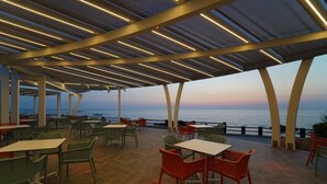 Zona pranzo all'aperto con tavoli e sedie, vista sul mare e un tramonto sullo sfondo.