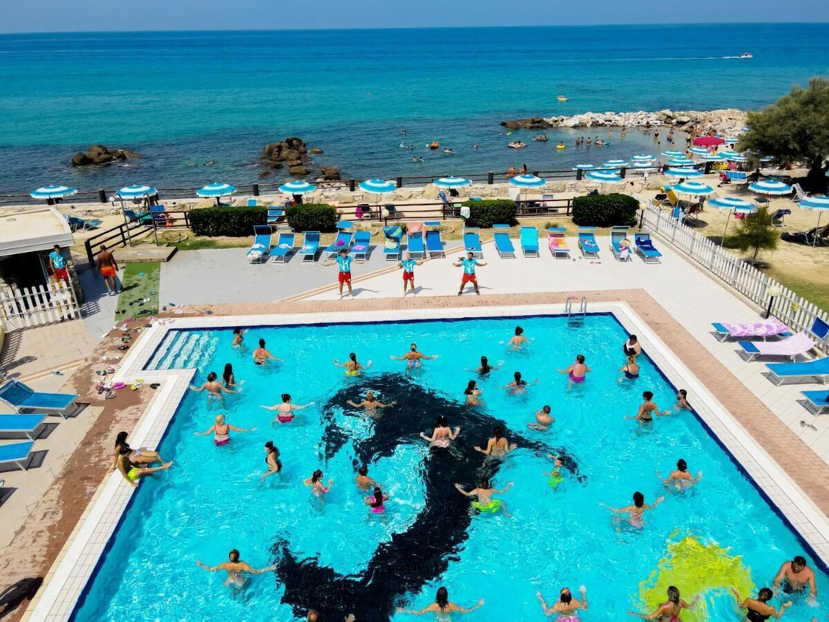 Ein Schwimmbecken mit Menschen im Wasser, umgeben von Liegestühlen und Sonnenschirmen, mit Blick auf das Meer.
