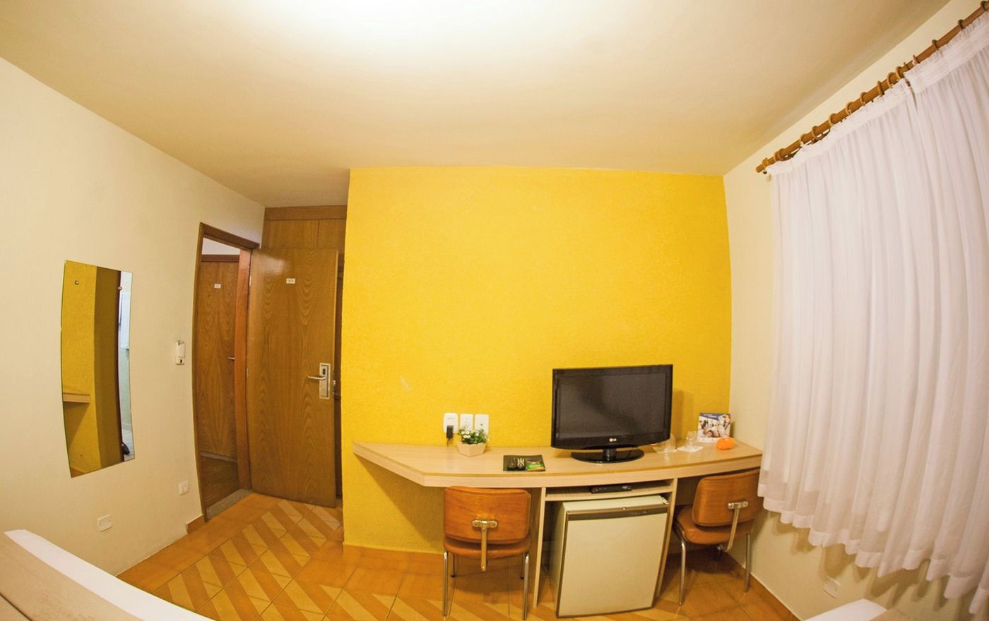 Quarto compacto com escrivaninha, televisão, mini-geladeira e uma cadeira.
