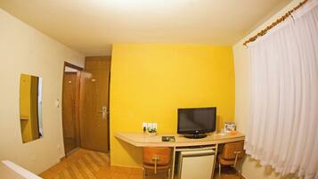 Quarto compacto com escrivaninha, televisão, mini-geladeira e uma cadeira.