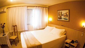 Habitación de hotel con una cama grande, una mesita de noche con lámpara, una silla, una pequeña mesa con un florero y un cuadro en la pared.