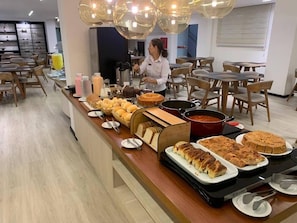 Montaje de bufé con diferentes opciones de comida, una persona sirviendo, y un interior moderno con muebles de madera e iluminación colgante.