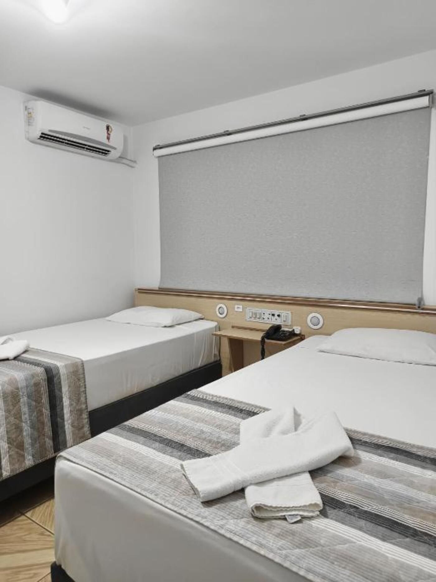 Quarto de hotel com duas camas, uma janela com persiana, um ar condicionado e uma mesa de cabeceira com telefone.