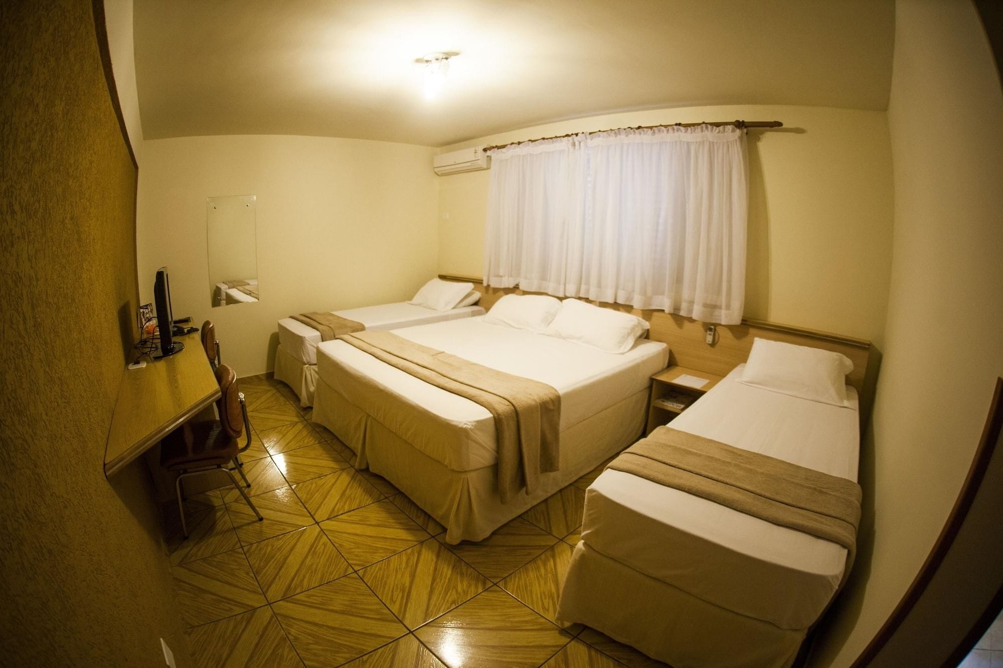 Quarto de hotel com duas camas, piso de madeira, escrivaninha com cadeira e janela com cortinas.