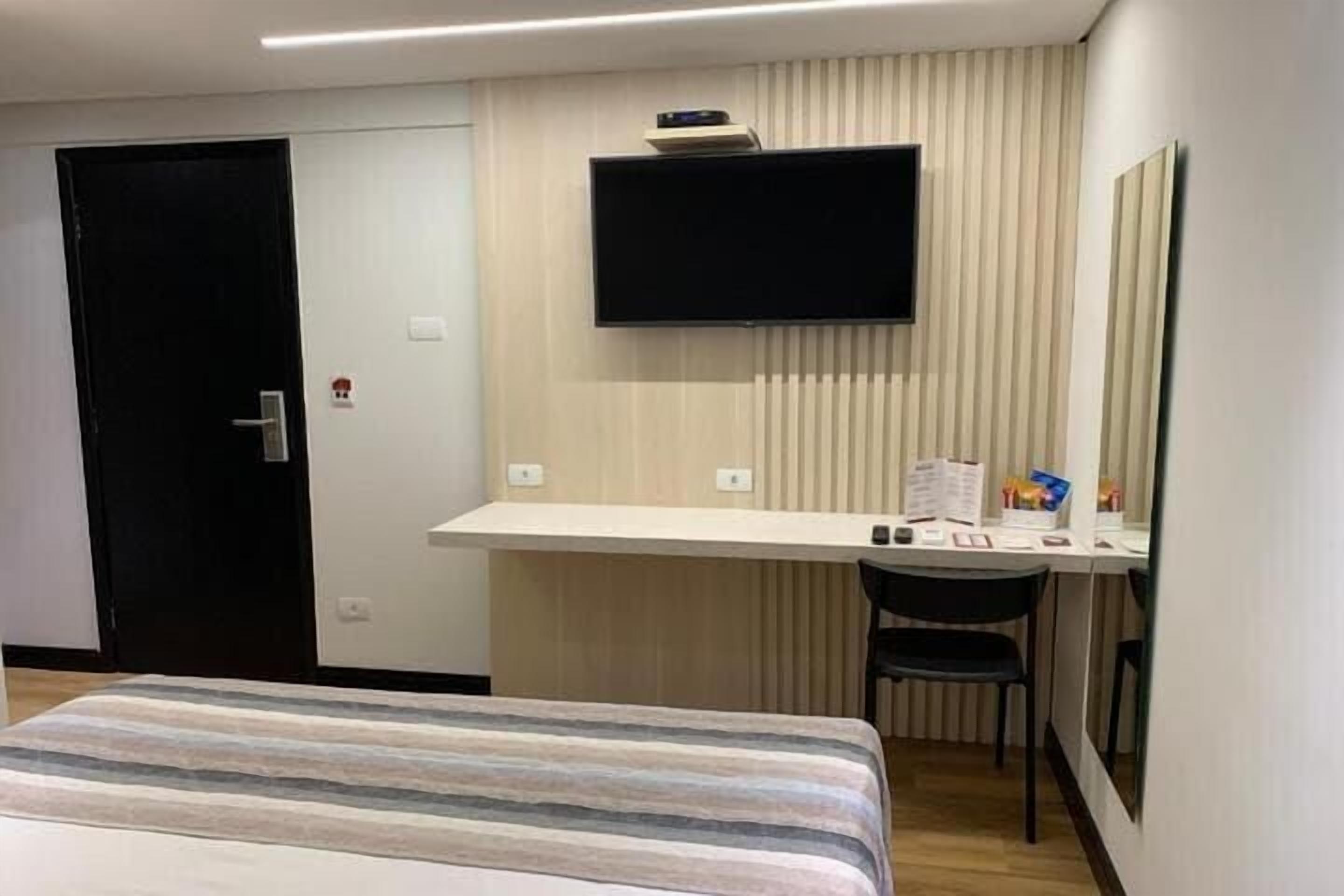 A hotel room with a bed, a desk, a chair, a flat-screen TV, and a door.