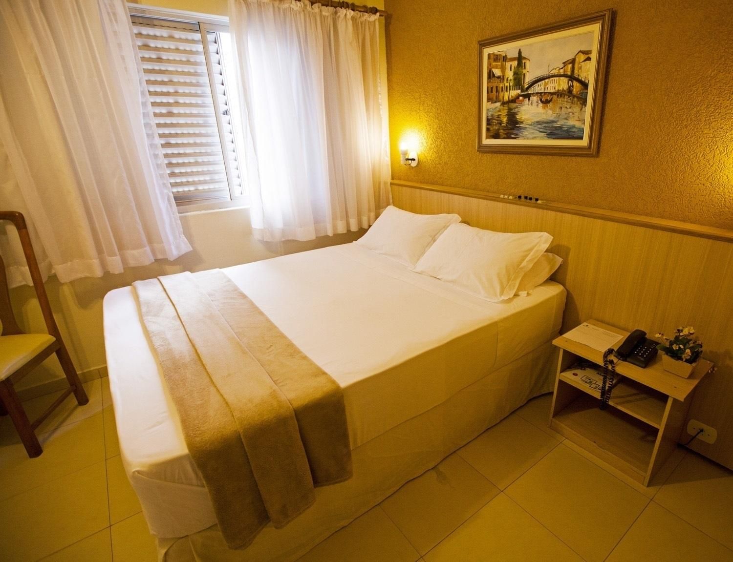 Quarto de hotel com cama, uma cadeira, mesa de cabeceira com telefone e um quadro na parede.