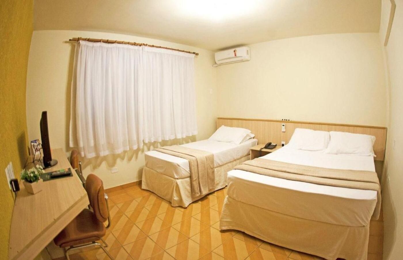 Quarto de hotel com duas camas, uma mesa com cadeira, uma janela com cortinas e um aparelho de ar condicionado.