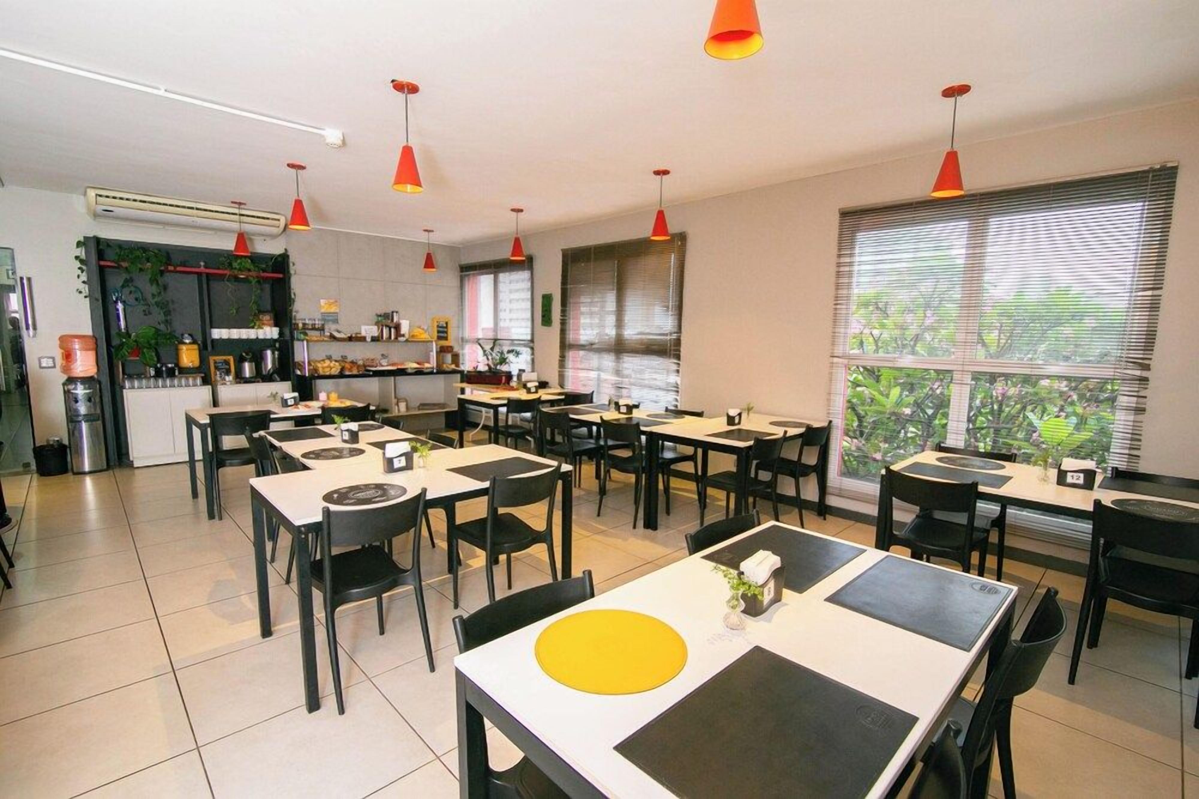 A spacious dining area with multiple tables and chairs, a kitchen area with shelves and appliances, and large windows with blinds.