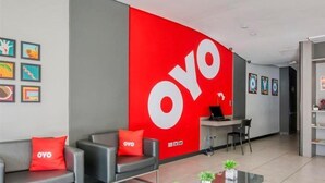 Un espacio de oficina moderno con una gran pared roja que presenta el logotipo 'OYO', un mostrador de recepción y una zona de estar con dos sofás y una mesa de centro.